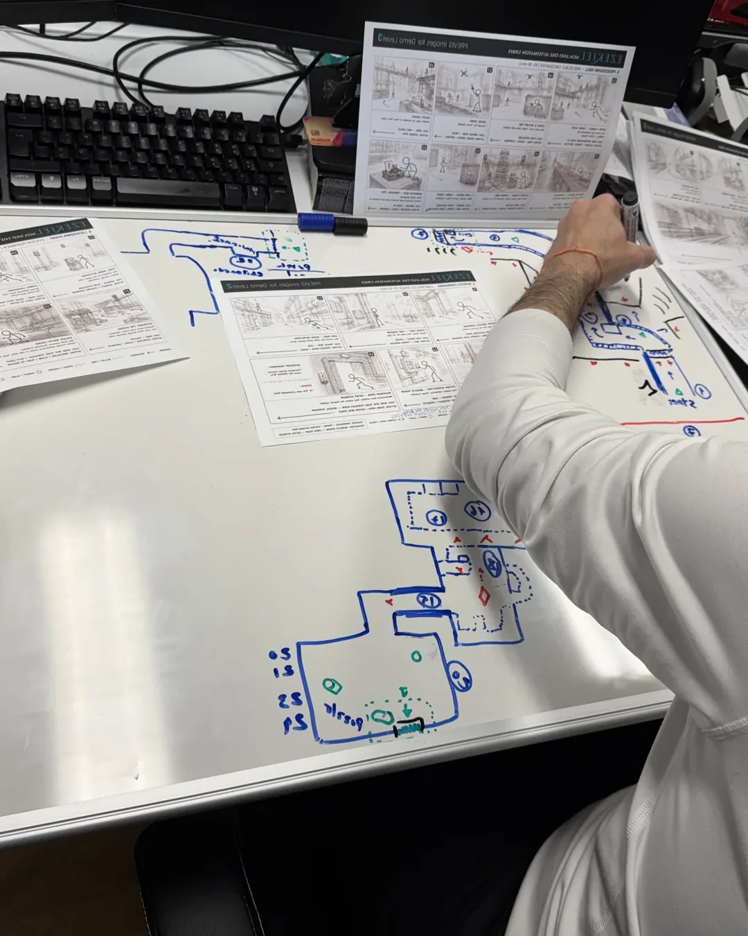 An NC Gaming designer sketching an Ezekiel level layout in marker on a whiteboard desk, printed design sheets spread around it.