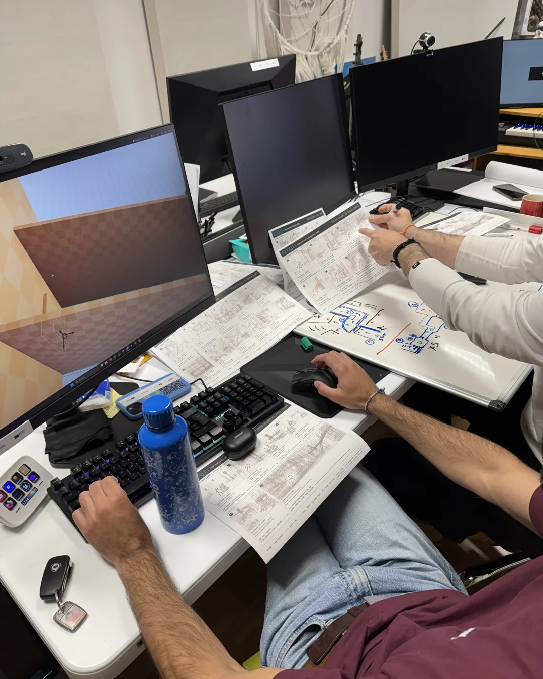 Two NC Gaming developers at a desk working through Ezekiel previs sheets and a hand-drawn level map, a 3D editor open on the monitor.